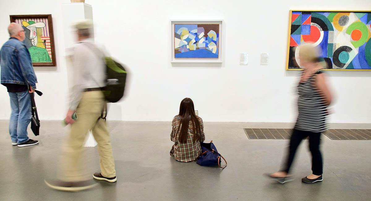 People in an art gallery, one young woman sitting on the floor in front of a painting while others walk past