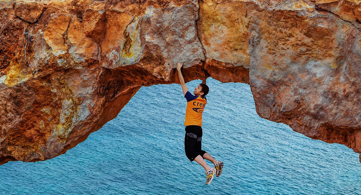 Man hanging from the bottom of a natural stone bridge over the sea
