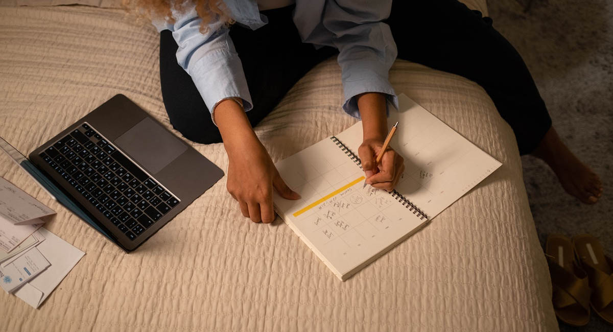 close-up woman's hands writing on calendar on bed with laptop