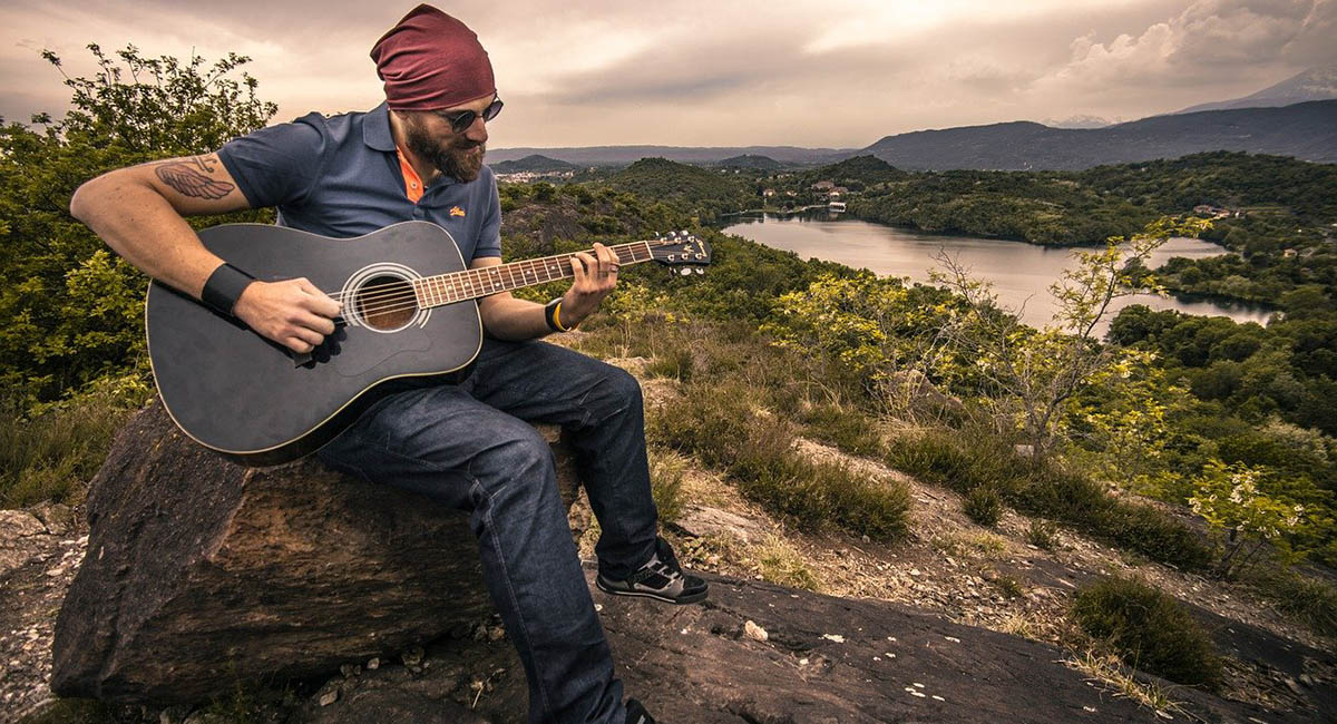 White man playing guitar on hill by small lake
