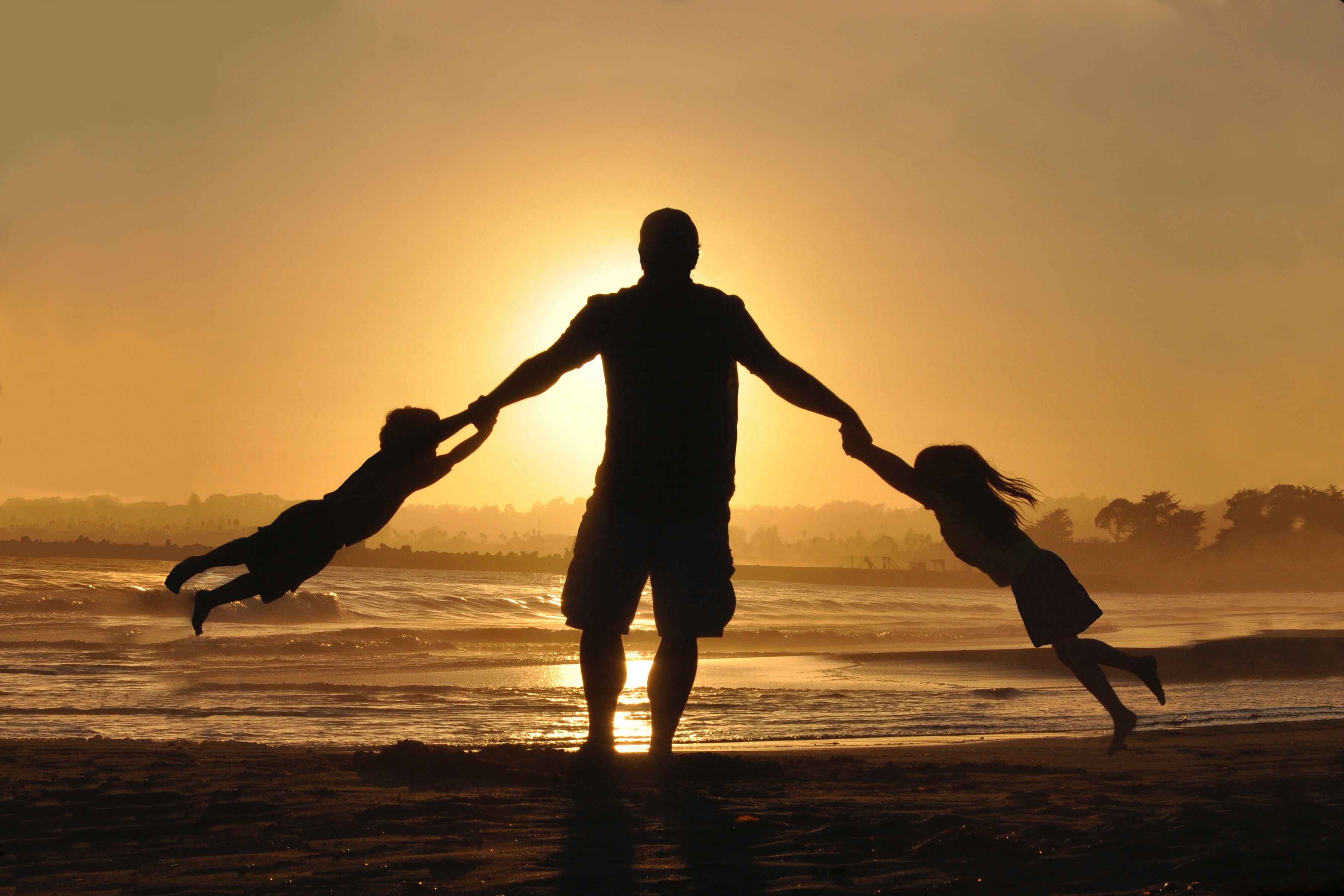 Silhouette of a man swinging two children by the hands on a beach