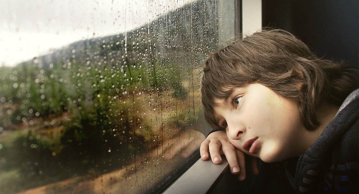 child looking out train window in the rain with a sad expression