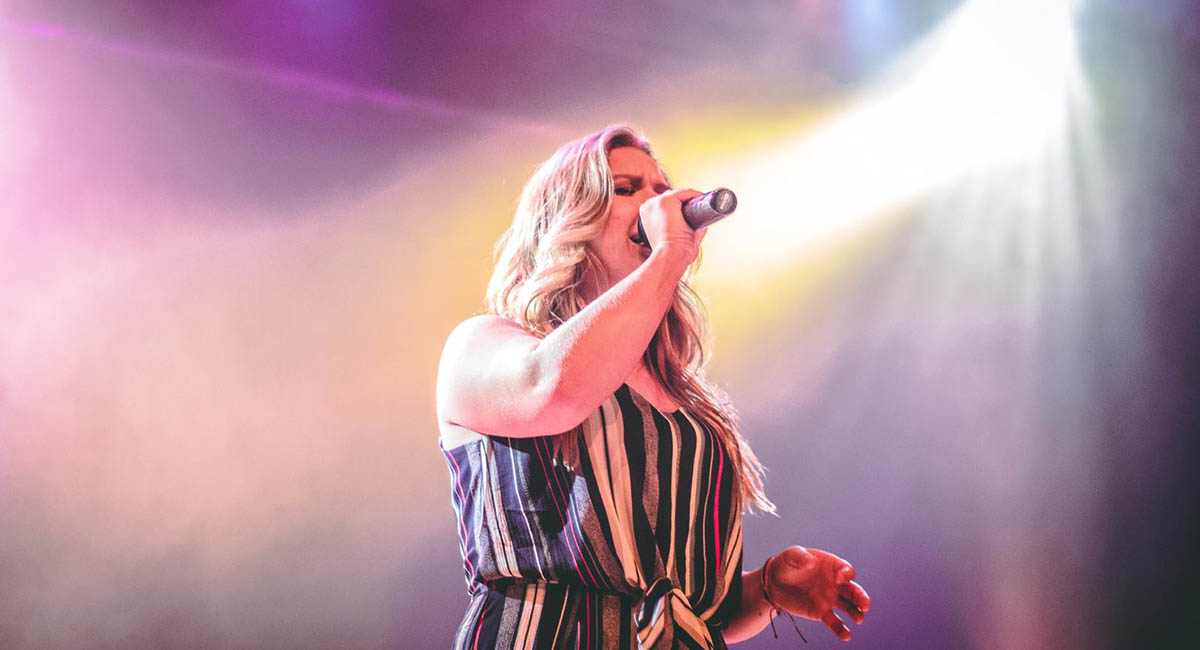 White woman with blond hair and striped dress singing on stage with spotlight behind her
