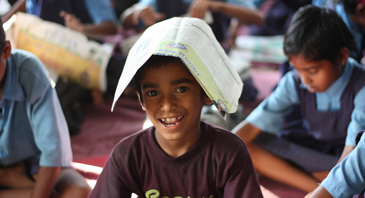 Young boy in school smiling with newspaper over his head, students at desk in background, India