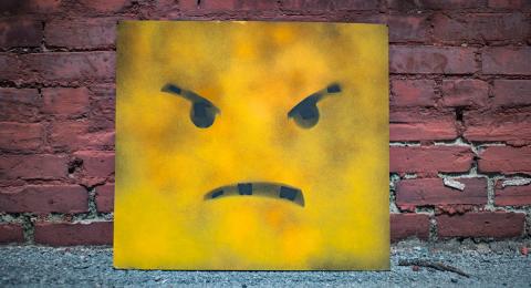 Yellow square with angry face leaning against a brick wall