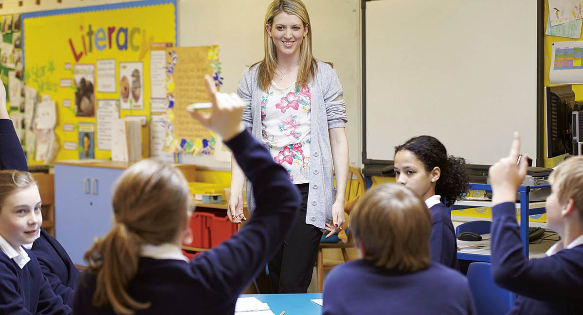 female white teacher standing with smile in front of seated students with hands raised